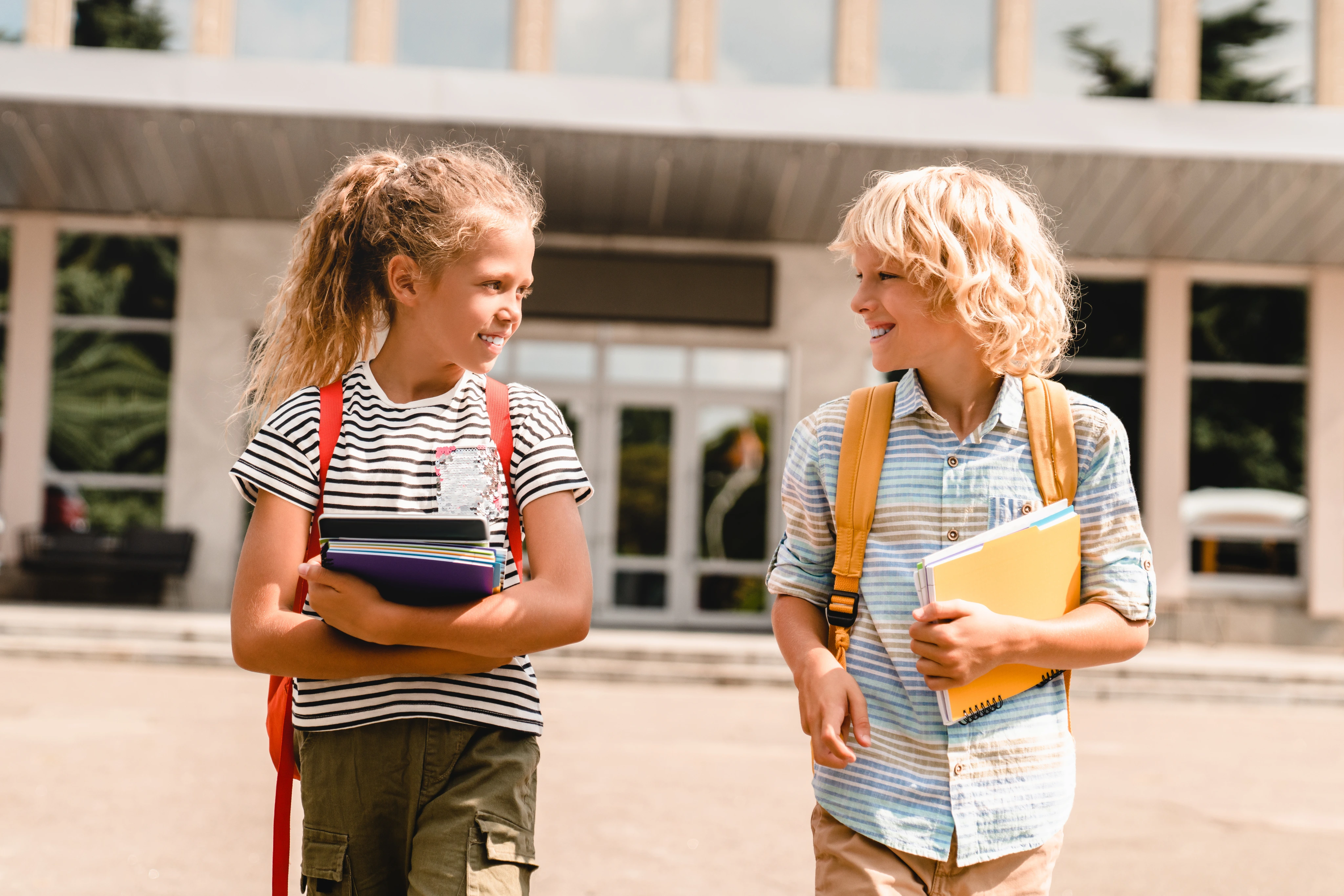Duas crianças caminham com livros e mochilas na escola.